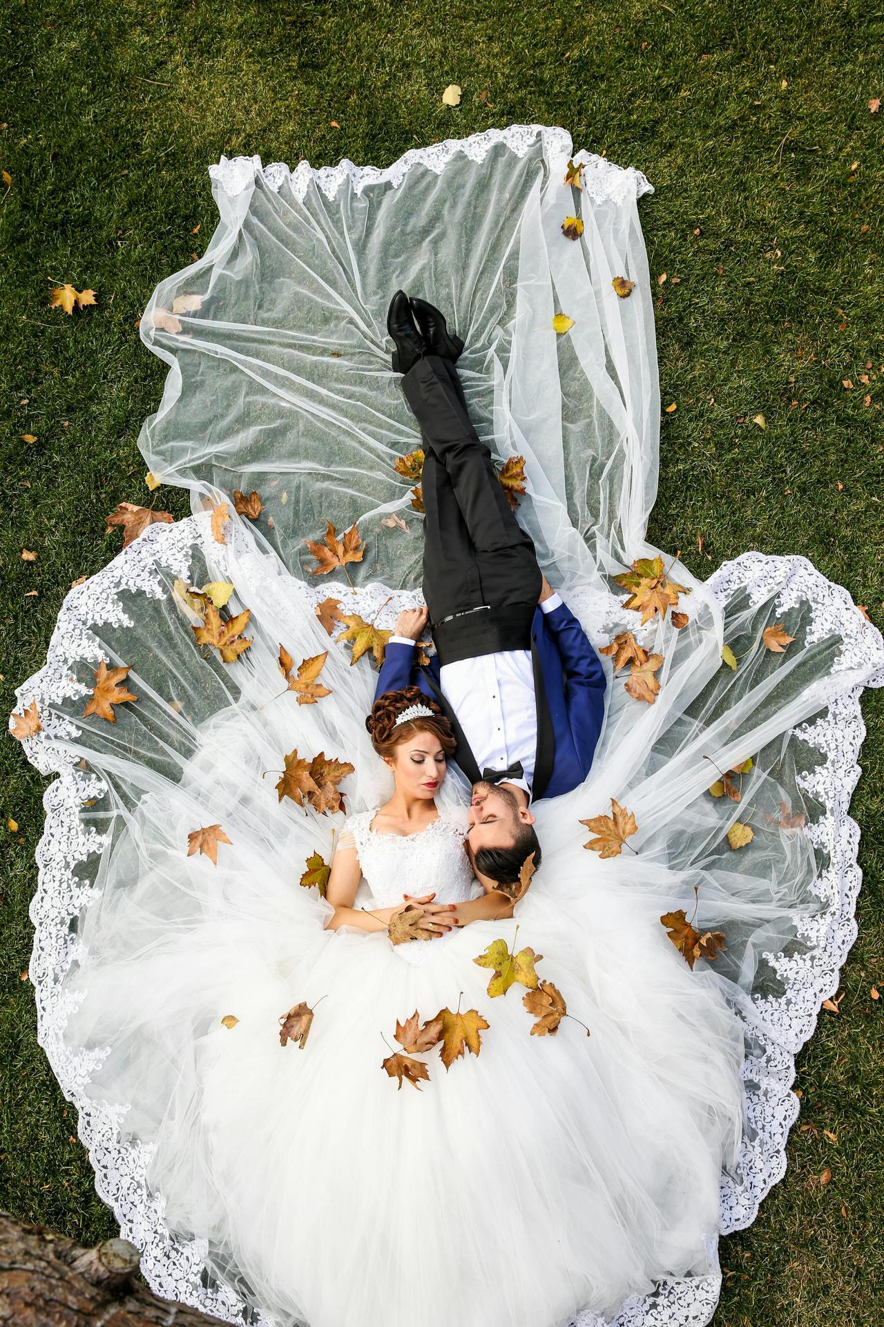 Bride and groom lying together on a lawn surrounded by autumn leaves, captured from above.